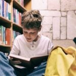 Boy sits on floor between library shelves, reading a book with interest