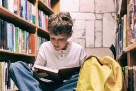 Boy sits on floor between library shelves, reading a book with interest