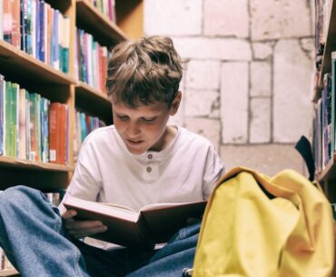 Boy sits on floor between library shelves, reading a book with interest