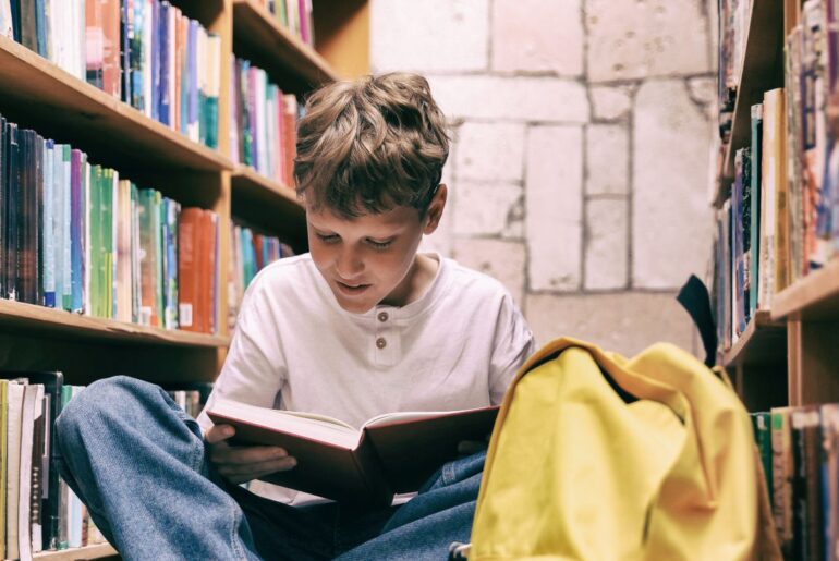 Boy sits on floor between library shelves, reading a book with interest